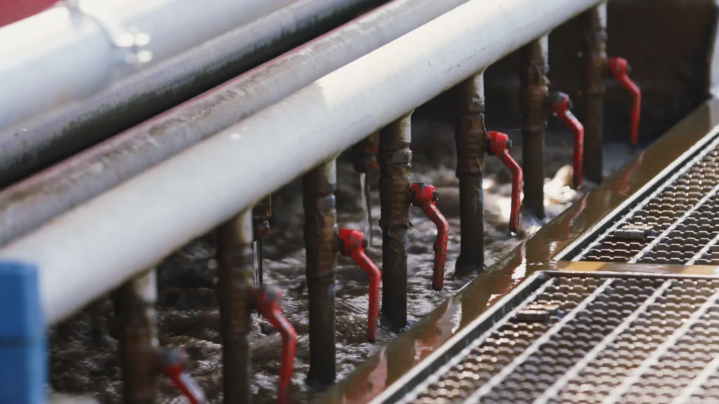 Close-up of white water pipes with red ball valve handles over a trough of churning brown water beside a metal grate.