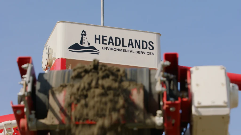 Headlands Environmental Services logo on a white tank above a blurred conveyor carrying damp sand, under a clear blue sky.