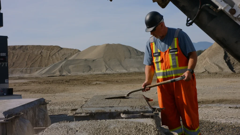 A man in orange overalls shovels fresh concrete into a steel block mold, with sand stockpiles behind him.