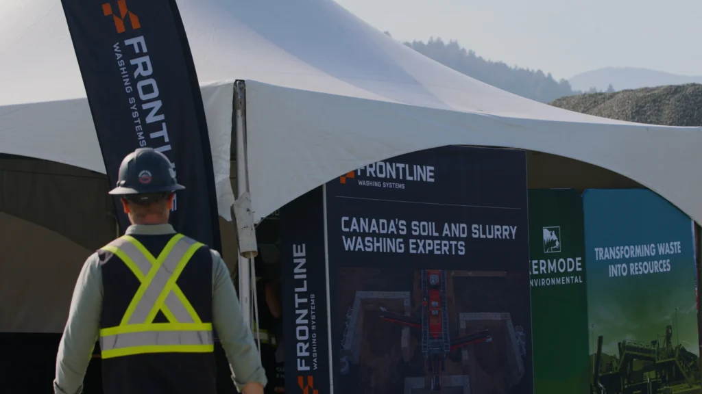A man in a hi-vis vest walks toward a white tent with Frontline and Kermode Environmental display panels and a Frontline banner.
