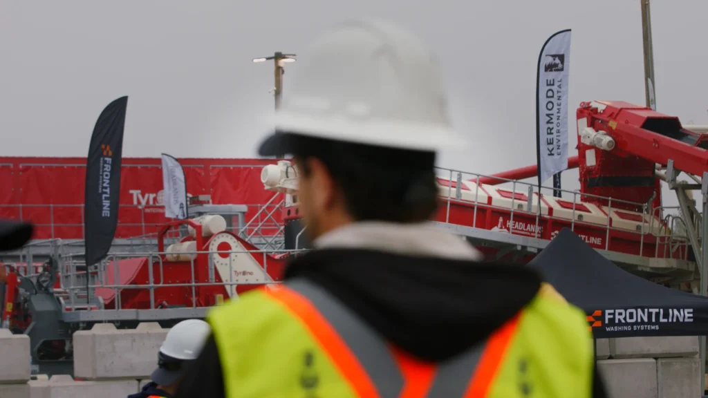 A blurred attendee in a white hard hat stands in the foreground, with the red wash plant and Frontline and Kermode Environmental flags behind.