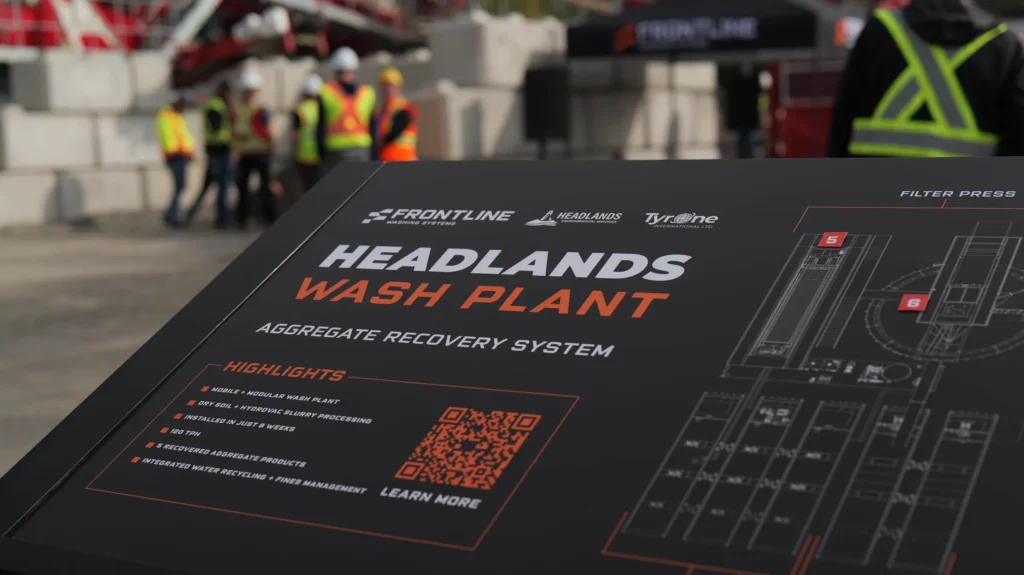 Black display board titled Headlands Wash Plant, listing highlights beside a QR code and a plant layout, with visitors blurred in the background.