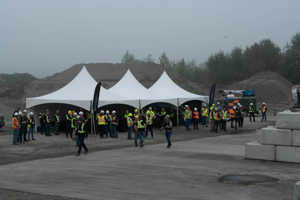 Attendees in hi-vis vests and hard hats stand under three white peaked tents on a foggy morning, with gravel stockpiles behind them.