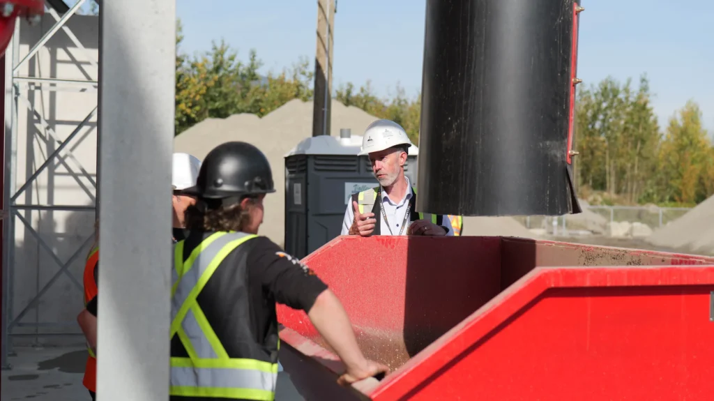 Mike Hofer speaks from behind a red hopper, with a large black pipe overhead and two listeners in the foreground.