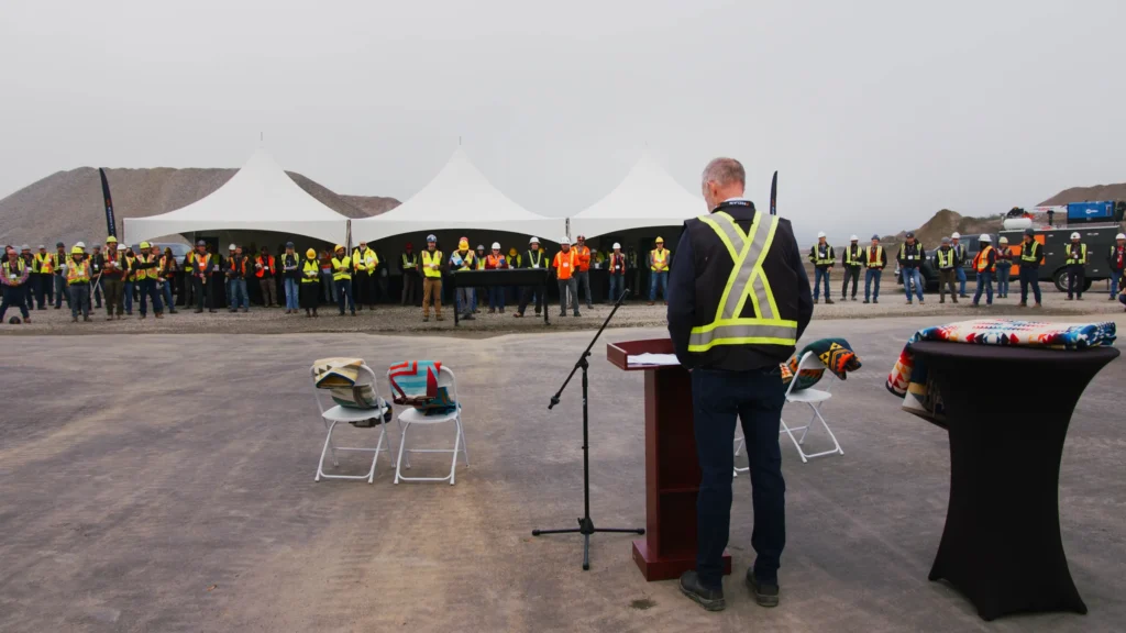 Mike Hofer at a wooden podium faces a crowd gathered around white tents, with patterned blankets draped over chairs and a table.