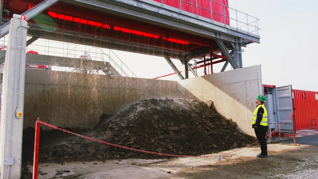 A man in a green hard hat looks up at a red filter press as dry cake falls onto a pile behind a chain barrier.
