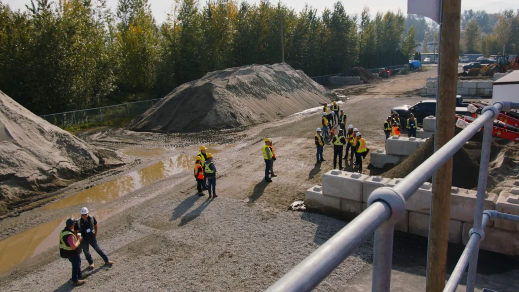 Looking down from a steel railing, small groups of attendees stand among sand stockpiles, puddles and concrete blocks.