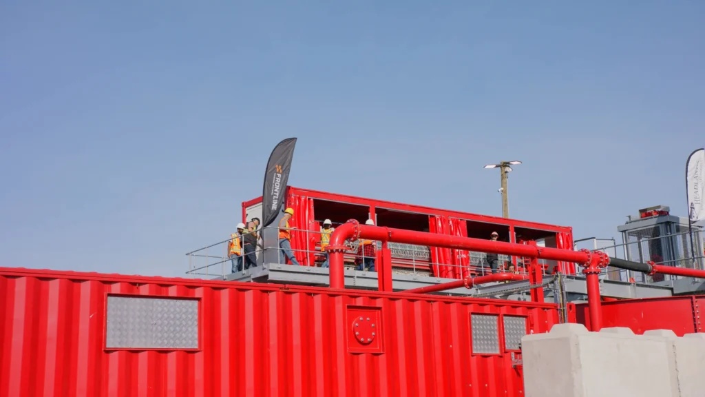 Attendees walk along a raised platform beside a Tyrone filter press container under a clear blue sky.