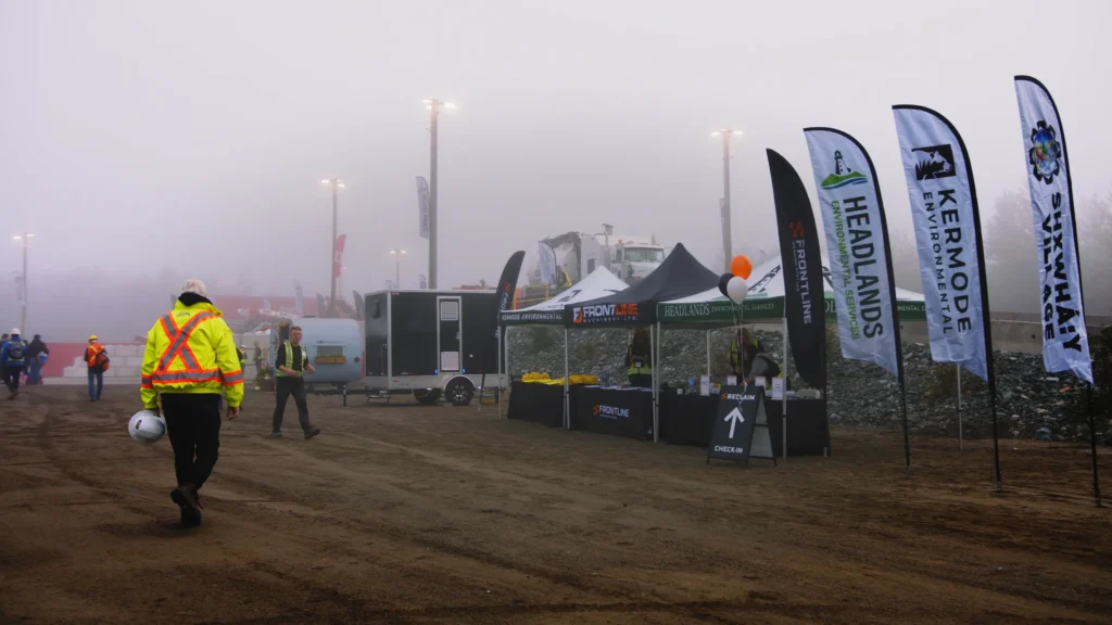 A man in a hi-vis jacket carries a white hard hat toward the check-in tents, lined by Frontline, Headlands, Kermode Environmental and Shxwhá:y Village flags.