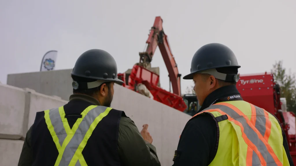 Two people in black hard hats and hi-vis vests watch a red excavator load material into a hopper beyond a concrete wall.