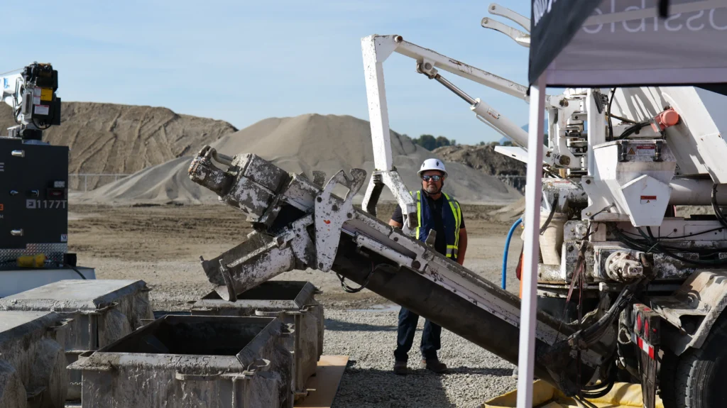 A man in a white hard hat stands beside a concrete truck chute pouring into steel block molds.