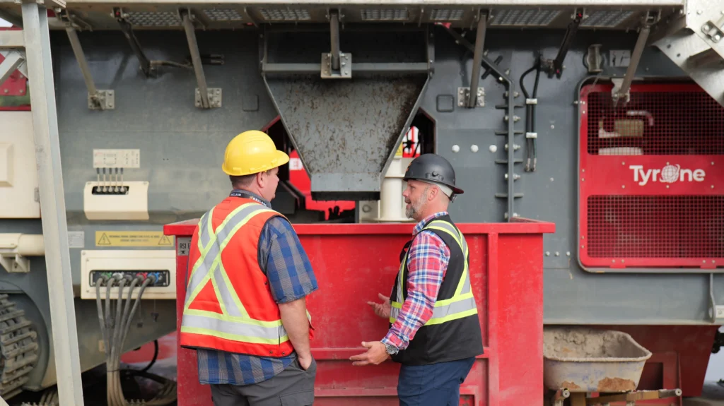 Two men in hi-vis vests talk in front of a red bin and the grey hopper.