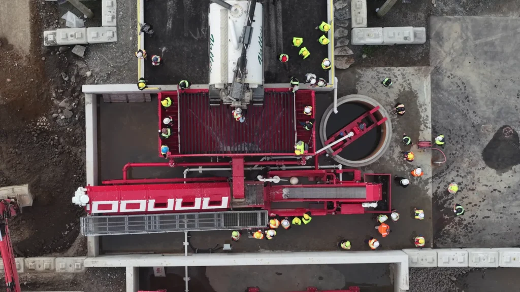 Top-down drone shot of the red wash plant, a vacuum truck over the grate, a round tank, and attendees gathered around the equipment.