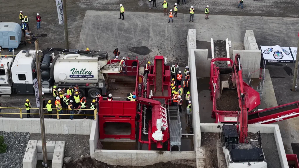 Angled drone view of a Valley Waste & Recycling truck discharging onto the red grate, with a crowd watching and an excavator feeding a second unit.